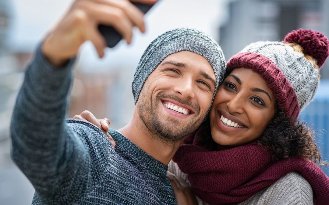 Happy couple taking selfie outdoors in winter clothing and beanies, smiling.