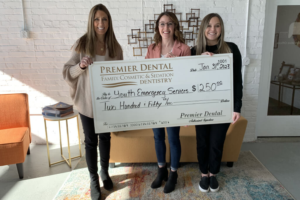 Three women holding large Premier Dental charity check in indoor setting.