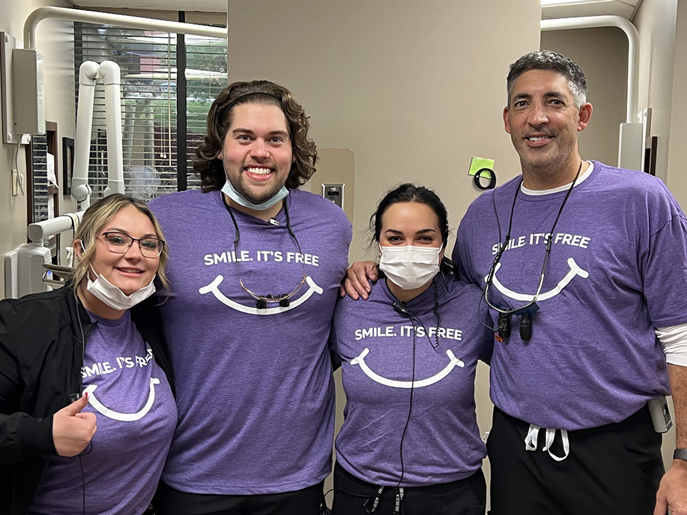 Four dental team members wearing purple shirts smile together in a modern dental office.