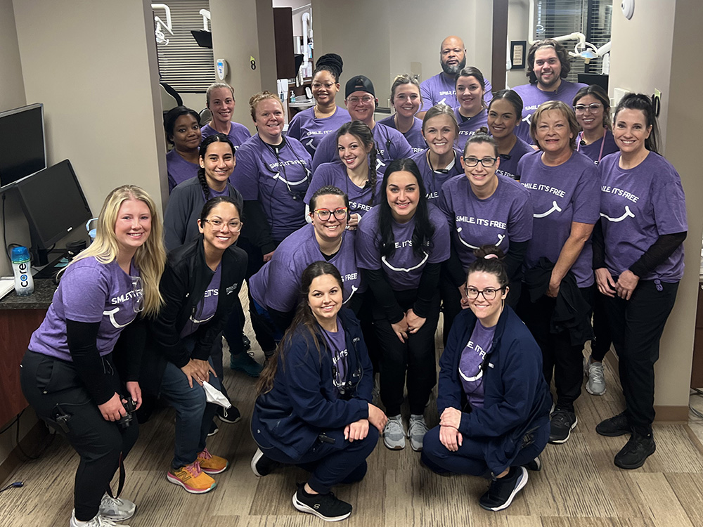 A diverse team of dental professionals in purple scrubs posing together in a clinical office setting.