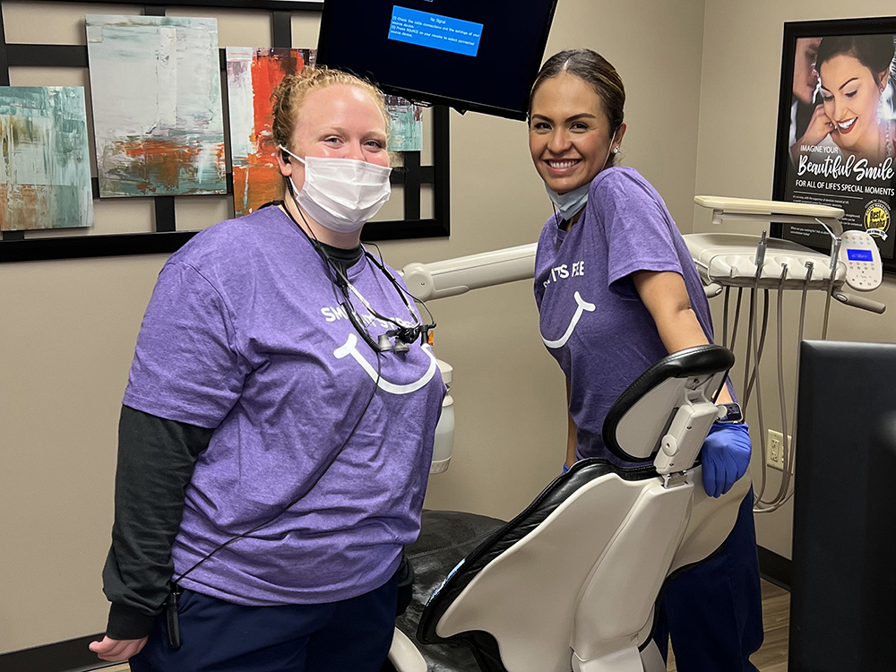 Two dental professionals in purple scrubs and masks work together at a dental treatment station in a clinical setting.