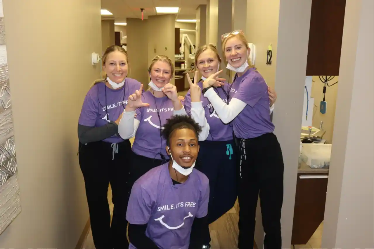Team of five dental professionals wearing purple scrubs posing together in clinic hallway.